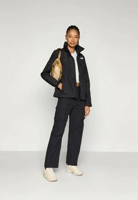 Mulher em pé a usar uma jaqueta preta, calças cargo pretas, ténis brancos, uma t-shirt curta branca e a carregar uma mochila bege num dos ombros.