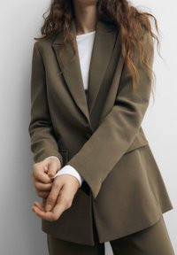 Blazer vert olive sur mesure avec une texture lisse, fermeture à un bouton et col cranté, associé à un haut blanc à manches longues.