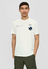 Biała bawełniana koszulka z ciemnoniebieskim wzorem kwiatowym po lewej stronie klatki piersiowej, z akcentami tekstowymi. Okrągły dekolt i krótkie rękawy, luźny krój.