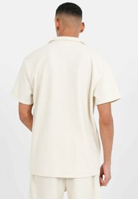 Camisa de manga corta de color crema con una tela de canalé texturizada y un cuello. La vista trasera muestra un corte relajado y un dobladillo recto.