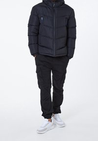 Uomo che indossa una giacca a vento nera, pantaloni cargo neri e sneakers bianche, in piedi con le mani nelle tasche su uno sfondo bianco.