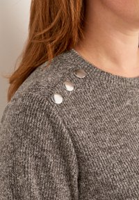 Grijze gebreide trui met een gestructureerd patroon, ronde halslijn en drie metalen knopen die de schouder accentueren.