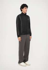 Chaqueta negra con cremallera y detalles de ribete blanco, combinada con pantalones de pierna ancha grises y zapatillas blancas. Tejido suave con un diseño ajustado.
