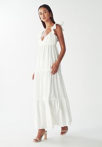 Valkoinen maksimekko, jossa on röyhelöolkaimet, syvä V-aukko ja kerroksellinen hame. Sileä kangas, jossa on virtaava muotoilu. Yhdistetty nauhakorkokenkiin.