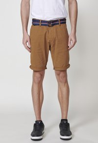 Braune Baumwollshorts mit umgekrempelten Säumen, kombiniert mit einem marineblauen und roten gestreiften Gürtel. Getragen mit schwarzen Sneakers.