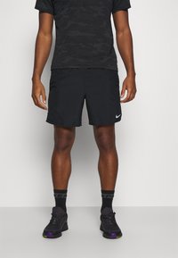 Mustat Nike-shortsit, kevyet, valkoisella swoosh-logolla. Yhdistetty mustaan, kamouflagekuvioiseen paitaan ja mustiin urheilukenkiin violeteilla yksityiskohdilla.
