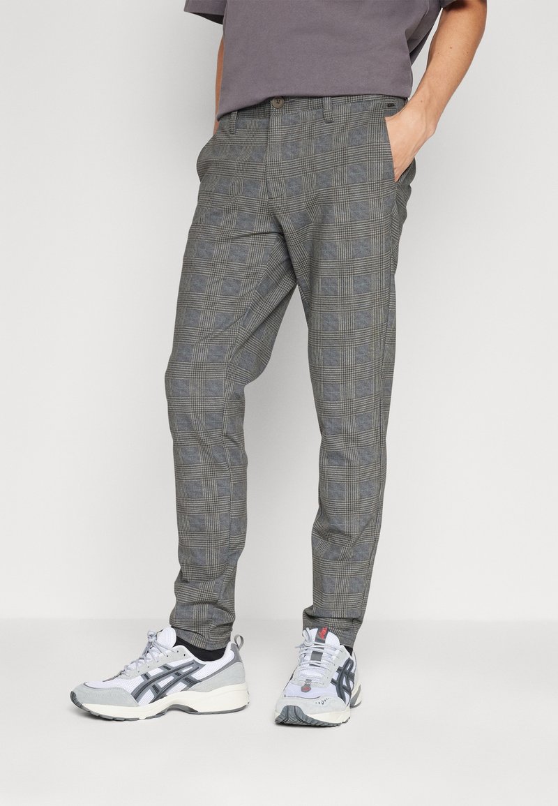 Pantalon à carreaux gris avec une coupe ajustée, présentant un motif à damier, des poches latérales, porté avec des chaussures de sport blanches et grises.