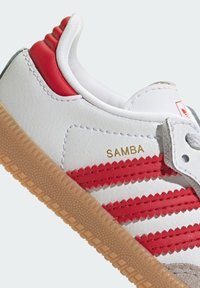 White sneaker with red stripes, gold "SAMBA" lettering, and gum sole. Features a padded collar and textured leather upper.