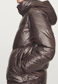 Brązowa kurtka puchowa o błyszczącym wykończeniu, z wysokim kołnierzem, przymocowanym kapturem i pikowaną fakturą. Widać boczne kieszenie.