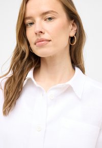 Biała koszula na guziki, wykonana z lekkiego materiału, z kieszenią. Posiada klasyczny kołnierzyk i luźny krój. Modelka ma średnio brązowe włosy i kolczyki w kształcie kół.