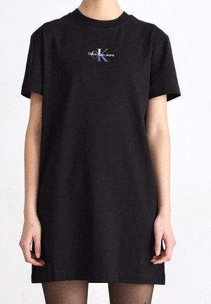 Osoba nosząca czarną sukienkę-t-shirt Calvin Klein Jeans z krótkimi rękawami i logo na środku klatki piersiowej na białym tle.