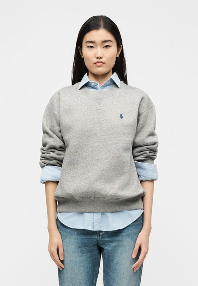 Νεαρή γυναίκα με γκρι φούτερ πάνω από ένα ανοιχτό γαλάζιο πουκάμισο με σηκωμένα μανίκια και μπλε τζιν, που στέκεται μπροστά σε ένα απλό φόντο.