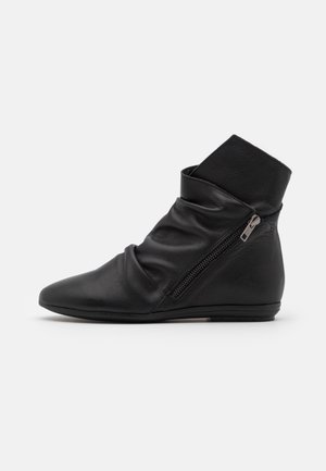 Bottine en cuir noir avec fermeture éclair latérale, texture légèrement froissée, semelle plate et bout pointu, vue de profil sur fond blanc.