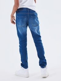 Jeans di denim blu con vestibilità slim, texture sbiadita, cintura elasticizzata, due tasche posteriori, abbinati a sneakers bianche.