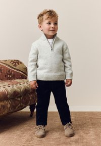 Lysegrå strikket trøje med halvlynlås i kraven, mørke bukser og beige sko på et barn. Baggrunden viser et vævet tæppe og en mønstret sofa.