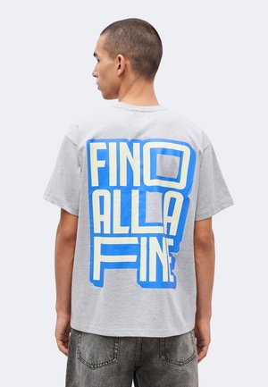 Młody mężczyzna z krótkimi włosami, ubrany w szarą koszulkę z dużym niebiesko-białym napisem „Fino Alla Fine” na plecach, stoi na jednolitym tle.