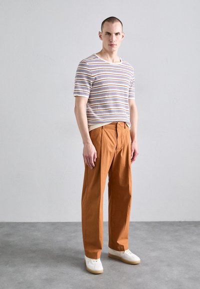 Chemise tricotée à manches courtes rayée en blanc, bleu et orange, associée à un pantalon large couleur rouille. Finition avec des baskets blanches.