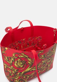 Czerwona torba tote z złotymi kwiatowymi wzorami, z podwójnymi uchwytami, wewnętrzną kieszenią na zamek i wyraźnym napisem "Versace Jeans Couture" w środku.