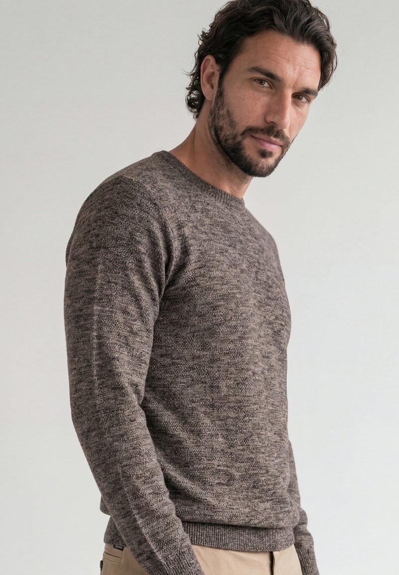 Homme aux cheveux foncés et barbe portant un pull marron texturé et un pantalon beige, légèrement de profil devant un fond clair uni.