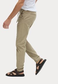 Beige lichtgewicht broek met een elastische tailleband en zijzakken, gecombineerd met donkerbruine sandaal met bandjes en een zwarte zool.