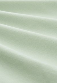 Gaiši zaļa auduma virsma ar gludu tekstūru, attēlo maigus krokas un maigu spīdumu, uzsverot tā vieglo un elpojošo īpašību.