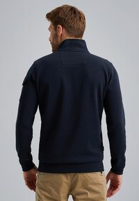 Suéter de color azul marino con un cuello alto, mangas largas y una textura lisa. Presenta un parche en la manga izquierda y un detalle bordado en la parte posterior.