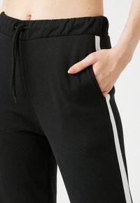 Pantalones de chándal negros con cintura elástica, cordón, bolsillo lateral y detalle de franjas blancas a lo largo de la costura exterior. Material suave de mezcla de algodón.