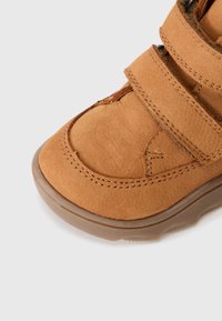 Close-up van een tan suède kinderschoen met twee klittenbandsluitingen en een bruine rubberen zool op een witte achtergrond.