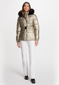 Veste à coussins dorée métallisée avec une capuche en fourrure noire amovible, une ceinture à la taille cintrée et des boutons sur le devant. Associée à un pantalon droit blanc.
