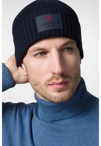 Granatowa dziergana czapka z teksturowanym wzorem ściągacza, z czarną łatą z logo i czerwonymi akcentami. Noszona z niebieskim swetrem z golfem.