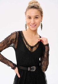 Zwarte kanten jurk met lange doorschijnende mouwen, V-hals, gegolfde randen en een zwarte riem met een zilveren gesp. Gedetailleerde kantstructuur overal.