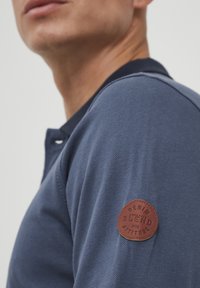 Námornícky modré polo tričko s textúrovanou tkaninou; na ľavom rukáve sa nachádza hnedá kožená nášivka s embosovaným textom "Denim Blend Attitude."