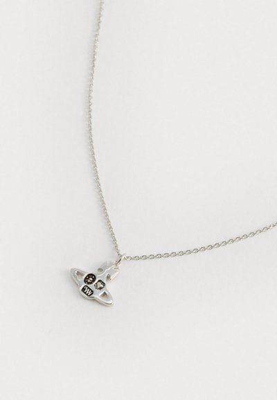 Corrente de prata com um pequeno pendente ornamentado, apresentando detalhes em preto e um design abstrato de figura coroada sobre um fundo branco.