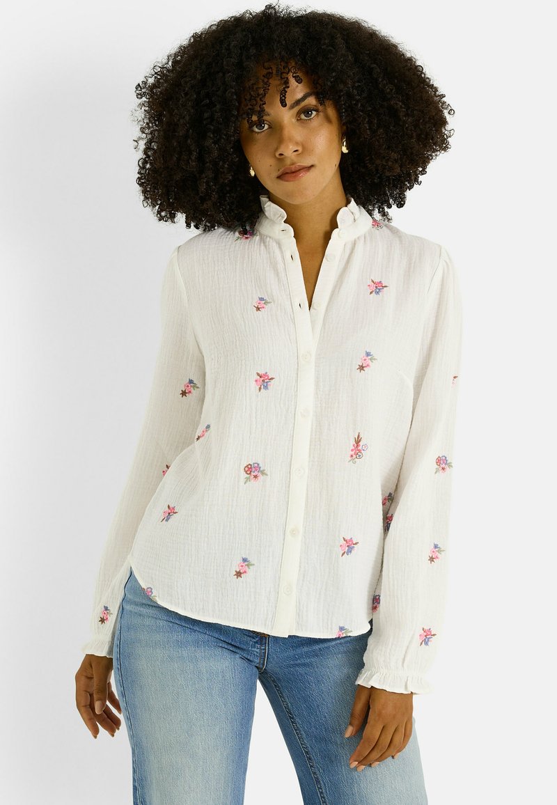 Camisa blanca abotonada con cuello con volantes y mangas largas, con bordados florales coloridos, confeccionada en tela texturizada.