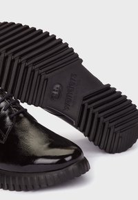 Zapatos de charol negro con diseño de punta afilada, que presentan una suela de caucho negro texturizada con estrías y el nombre de la marca "wonders" grabado.