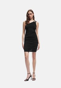 Robe noire à épaule unique avec fronces, en tissu extensible. Elle présente des bretelles décoratives et une silhouette ajustée. Associée à des talons hauts noirs.