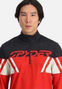 Maglietta a maniche lunghe rossa e nera con colletto alto, dotata di chiusura con zip. Presenta motivi angolari bianchi e rossi e il marchio "SPYDER" in evidenza.