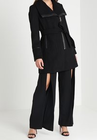 Cappotto nero con ampio revers, vita aderente e tasche con cerniera, abbinato a pantaloni neri con spacchi alti fino alla coscia e tacchi alti.