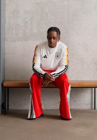 Mann i hvit langermet skjorte med svart og gul stripe og røde bukser sitter på en trebenk mot en betongvegg med hodet bøyd.