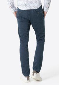 Dark blue cotton pants with a slim fit and two rear pockets featuring triangular flap closures. Worn with white sneakers.