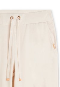 Gros plan sur la ceinture de pantalons de survêtement beige avec des cordons en tissu passant par des œillets dorés et une poche latérale avec une petite étiquette en métal.