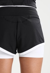 Black athletic shorts with a layered design over white inner shorts. Features a smooth texture and contrasting white trim along the hem.