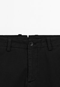 Pantaloni neri in tessuto texturizzato, con chiusura a bottone e passanti per cintura in vita. Tasche anteriori visibili.