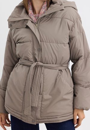 Kvinna som bär en gråbrun bältad puffjacka över en flerfärgad stickad tröja och mörkblå jeans, stående med avslappnade händer vid sidorna.