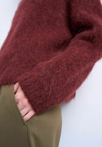 Personne portant un pull bordeaux duveteux avec la main partiellement dans la poche d'un pantalon vert olive, sur un fond clair uni.