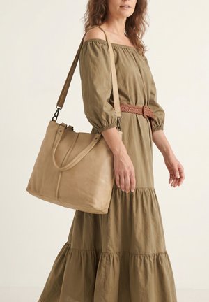 Frau in einem braunen schulterfreien Kleid mit geflochtenem Gürtel, die eine große beige Lederschultertasche am linken Arm trägt.