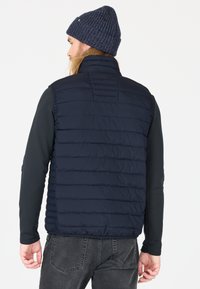 Marineblaues Pufferweste mit horizontalen Nähten, hohem Kragen und glatter Textur; getragen über einem schwarzen Langarmshirt, kombiniert mit einer gestrickten Mütze.