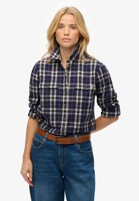 Navy and cream plaid flannel shirt with button front, roll-up sleeves, and two chest pockets, worn with high-waisted blue jeans and brown belt.