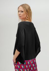 Černý pletený top s širokými rukávy; volný střih. Spárován s barevnými vzorovanými kalhotami s geometrickými tvary a živými barvami.