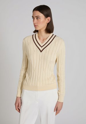 Donna che indossa un maglione lavorato a trecce color crema con strisce a V marroni e pantaloni bianchi, in piedi davanti a uno sfondo grigio semplice, che guarda a sinistra.